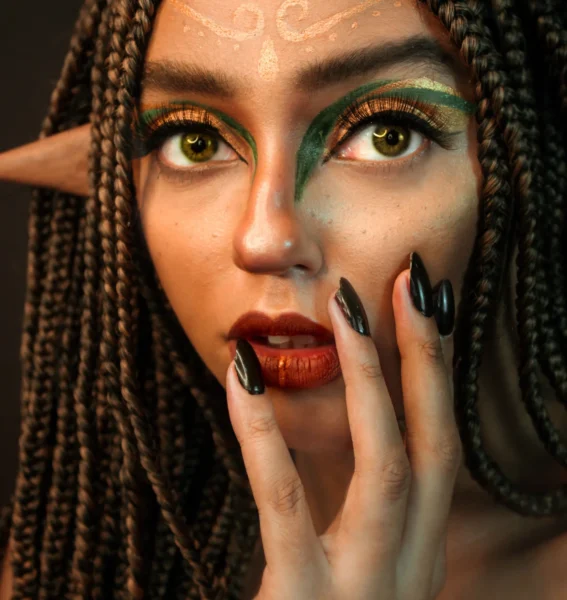 A woman with long braids, pointed elf ears, and golden swirled face paint gazing at the viewer.