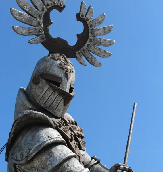A statue of an armored knight with a large winged crest on the helmet, holding a staff against a blue sky.